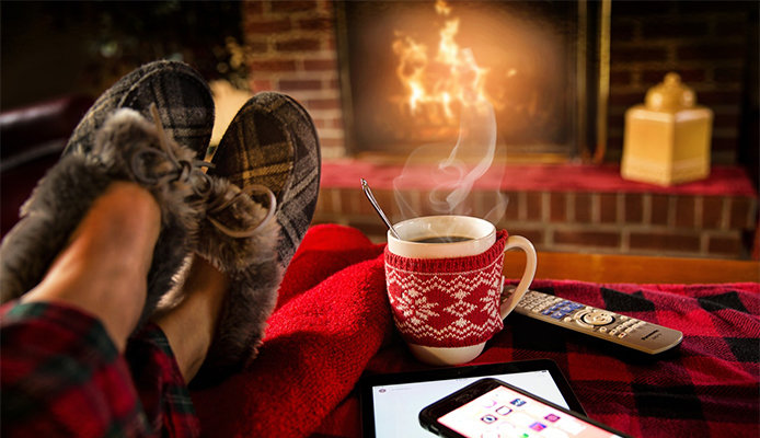 A person wearing grey moccasin slippers on a red plaid blanket is warming their feet by a brick fireplace. There's a steamy mug with a red winter pattern next to them, along with a tablet, a phone, and a television remote control.