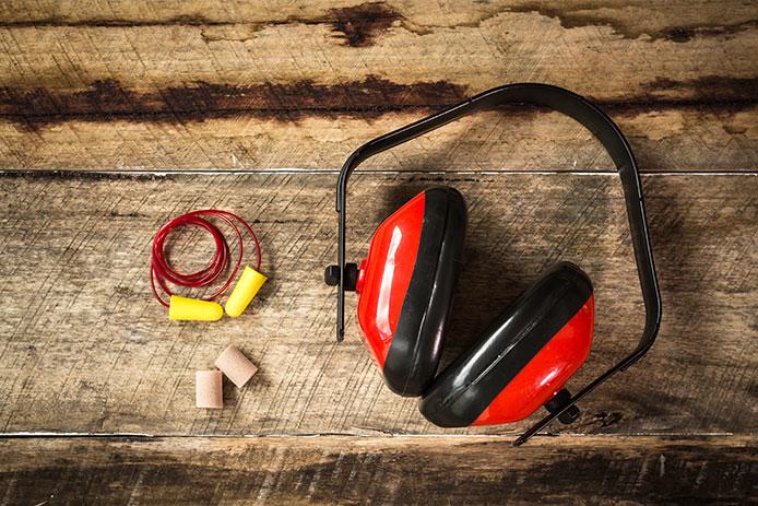 Several forms of ear protection are displayed against a wooden board. A headset with red and black earmuffs is featured next to two sets of foam earplugs, one with a cord in yellow and a flesh-toned cordless pair.