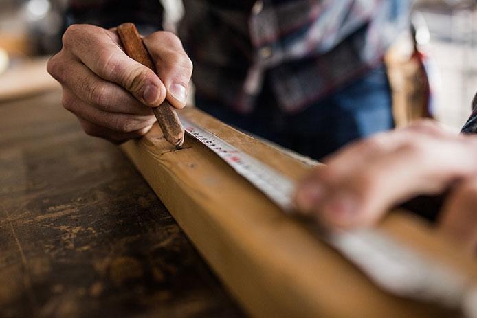 A person uses a white tape measure and a brown carpenter’s pencil to measure and mark up a long piece of wood.