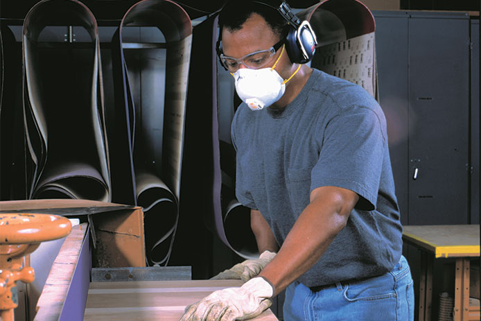 A man wearing a dark blue work shirt, jeans, a face mask, safety glasses, and construction earmuffs stands at a woodshop workstation, maneuvering a sheet of wood through a table saw with gloved hands.