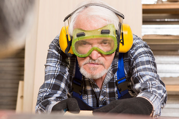 A man with white hair and a beard in a blue plaid shirt is wearing yellow earmuffs for protection and lime green safety goggles. He is looking at the surface of a piece of wood that he maneuvers with his hands protected by black work gloves.