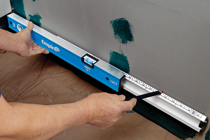 A person holds a blue and silver level with both hands as they press it against a wall and floor, using a pencil to mark the silver wall for their project.