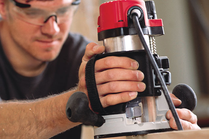 A man in safety goggles and a dark blue shirt holds a red and silver handheld wood router as he cuts into a piece of wood.