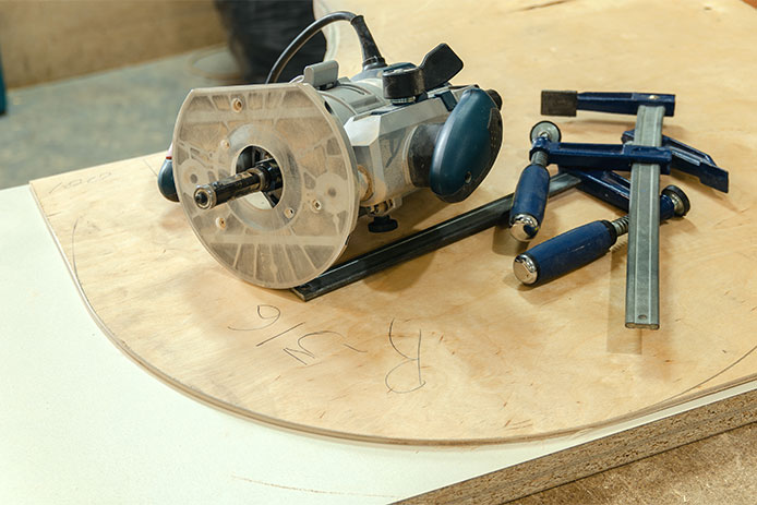 A silver-colored wood router rests atop a curved piece of light-colored wood. Several woodworking tools are piled next to the router.