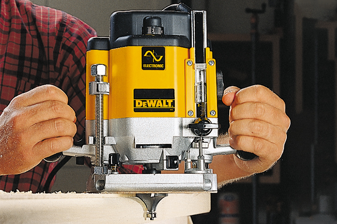 A person holds a bright yellow and black DeWALT brand wood router with both hands and cuts into a piece of light-colored wood.