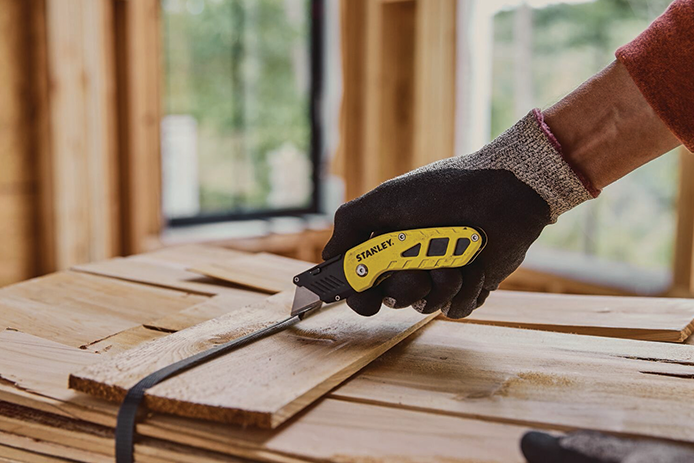 A person wearing a blue glove uses a yellow utility knife to cut the black strap binding a pile of boards together.