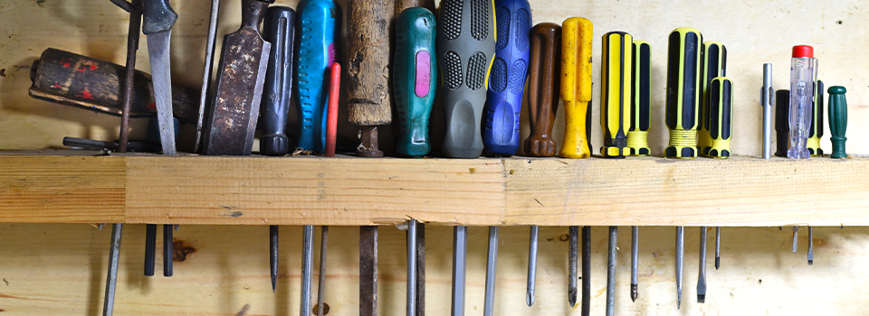 A multi-colored lineup of screwdrivers in various sizes and styles shows an example of basic homeowner tools. The screwdrivers are resting in a wall-mounted, wooden screwdriver holder.